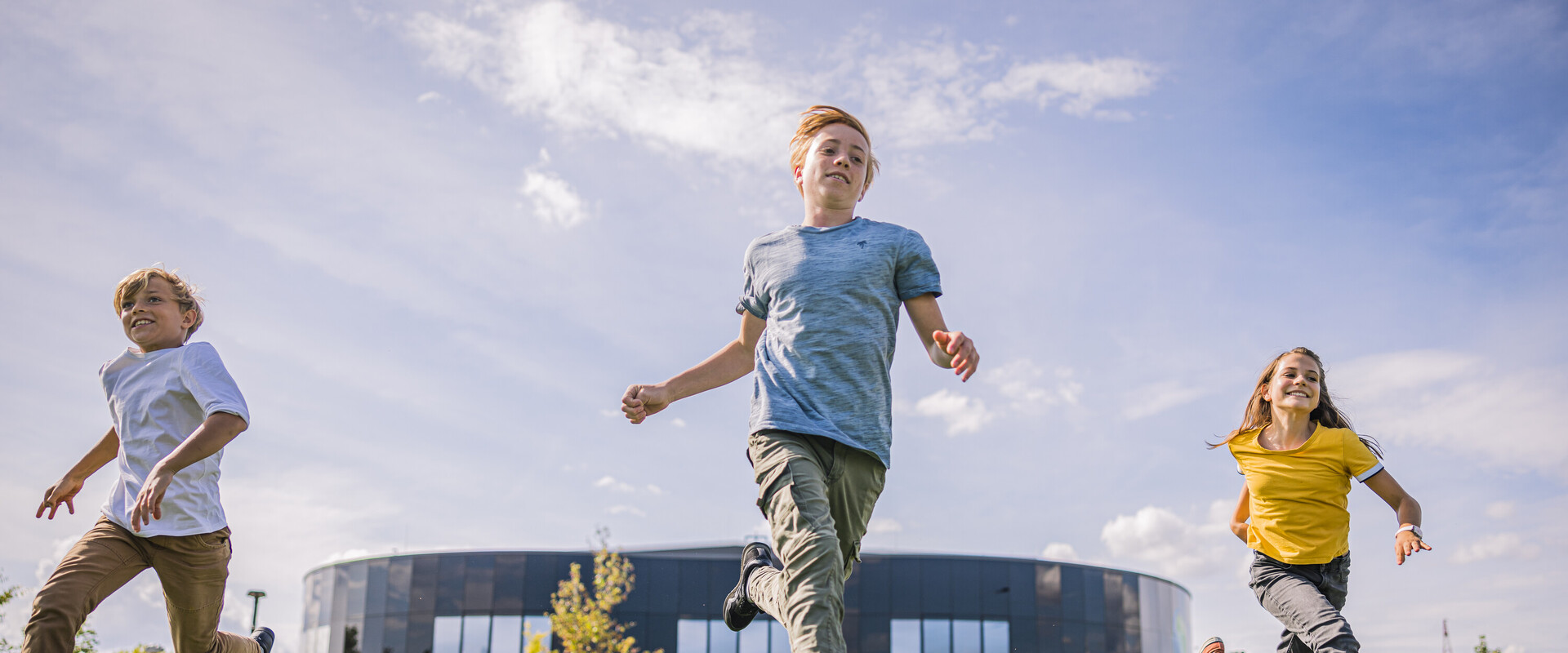 Drei Kinder rennen auf der Wiese im Au&szlig;enbereich, im Hintergrund sieht man das Geb&auml;ude der KLIMA ARENA.