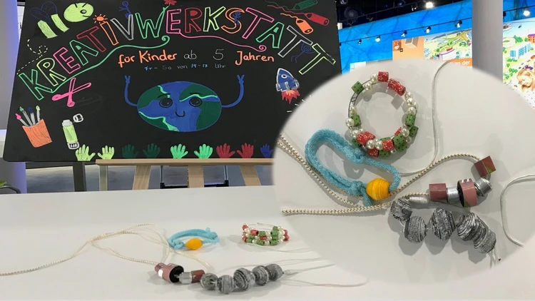 Auf dem Bild liegen gebastelte Papierperlenketten auf einem Tisch, im Hintergrund steht eine Tafel.