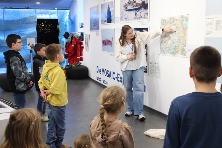 Eine Person erkl&auml;rt einer Gruppe von Kindern anhand einer gro&szlig;en Karte in einem Ausstellungs- oder Museumsraum.
